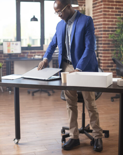 Young male employee with spectacles, carefully arranging his laptop at desk in brick wall workplace. Black businessman setting his digital device on table, preparing for work near office documents.