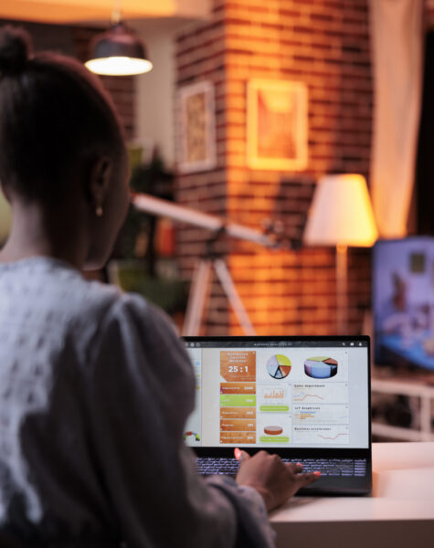 Female business analyst looking at diagrams and pie charts on laptop screen in modern home office with beautiful warm sunset light. Remote corporate worker analyzing company research result data